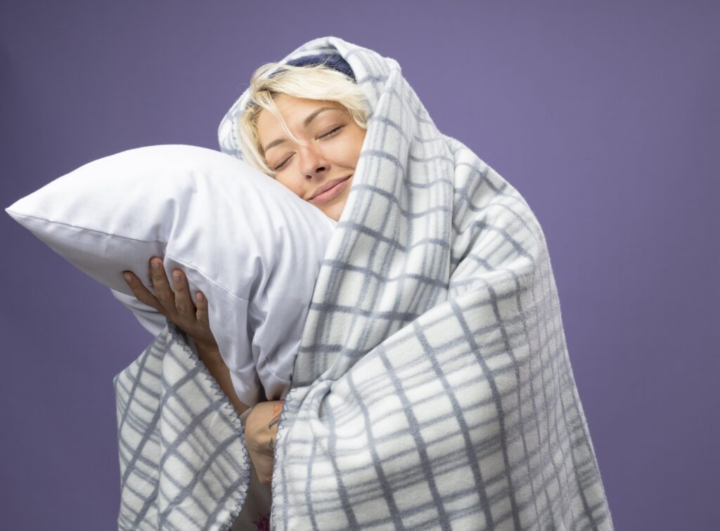 sick-unhealthy-woman-with-short-hair-warm-hat-wrapped-blanket-holding-pillow-leaning-her-head-pillow-with-closed-eyes-smilingover-purple-background